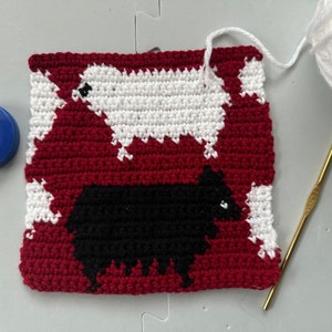 May include: A close-up of a crocheted square with two sheep, one black and one white, on a red background. The square is made with white, black, and red yarn. A crochet hook and a ball of white yarn are also visible in the image.