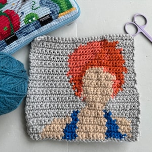 May include: A crocheted square featuring a stylized portrait of a person with orange hair, a blue and beige top, and a gray background.
