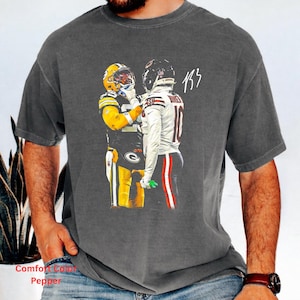 May include: A charcoal gray short-sleeve t-shirt featuring a graphic of two football players in uniform facing each other. One player wears a yellow and green jersey, while the other wears a white jersey with red accents. The text 133 is printed above.