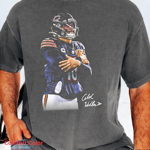 May include: A charcoal gray short-sleeve t-shirt featuring a graphic print of a football player in a navy blue uniform with a helmet, white pants, and orange accents. The graphic includes a white signature and the text Comfort Color pepper.