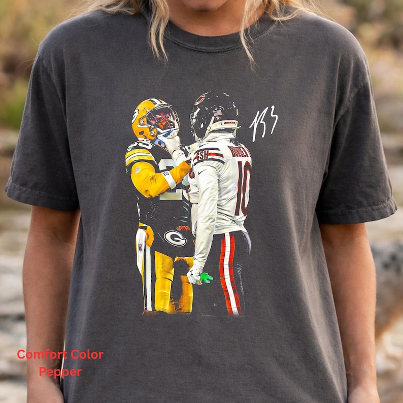 May include: A charcoal gray short-sleeve t-shirt featuring a graphic print of two football players in uniform facing each other. One player wears a yellow and green uniform, while the other wears a white uniform with red accents and the number 10.