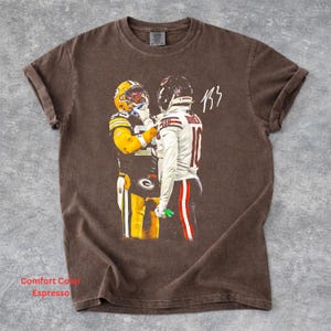 May include: A brown short-sleeve t-shirt featuring a graphic print of two football players in opposing uniforms facing each other. One player wears a yellow and green uniform, while the other wears a white uniform with red accents and the number 10.