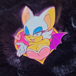 May include: A colorful enamel pin featuring a cartoon bat character with white, yellow, pink, and purple accents. The bat is set against a pink heart-shaped background with a gold border, and has glittery blue eyes.