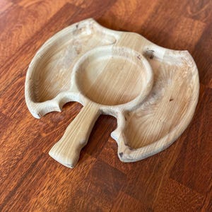 Puede incluir: Una bandeja de madera clara con asa y dos secciones. La bandeja tiene un patrón de vetas de madera natural y está diseñada con una forma orgánica única. El asa está unida a una sección redonda, y la otra sección tiene forma de hoja.