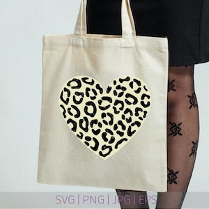 May include: A beige canvas tote bag featuring a large, centered heart graphic. The heart is filled with a black and cream leopard print pattern. The bottom of the image displays the text SVG | PNG | JPG | EPS in a light purple font color.