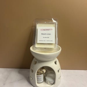 May include: A white ceramic essential oil warmer with heart-shaped cutouts. A clear plastic package of soy wax melt labeled "Beach Linen" sits on top. A small jar and a candle are inside the warmer.