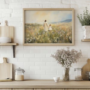 May include: A painting depicting a figure in a white robe walking with a child through a field of wildflowers. The painting is framed in a light wood frame and is hanging on a white brick wall.