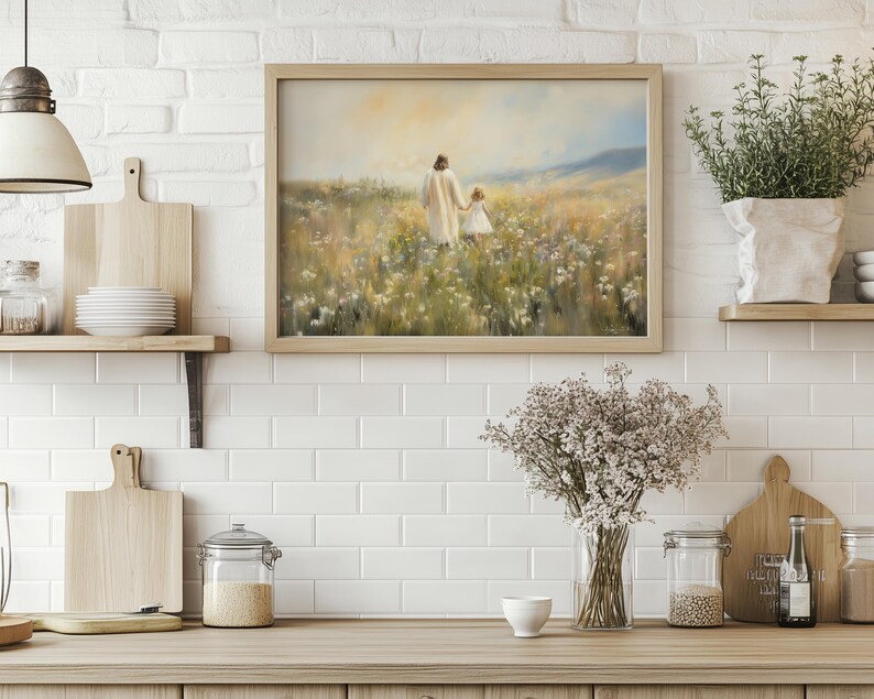 May include: A painting depicting a figure in a white robe walking with a child through a field of wildflowers. The painting is framed in a light wood frame and is hanging on a white brick wall.