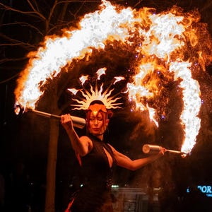 Pode incluir: Um artista com um vestido preto de forro vermelho e um adorno de cabeça em forma de sol segura duas tochas acesas, criando um grande arco de fogo contra um fundo noturno. O artista está num palco com pequenas chamas no chão.