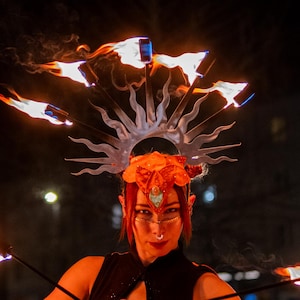 May include: A performer wearing a silver sunburst headpiece with orange floral accents and a black sleeveless top, holding two thin metal rods with bright orange flames at the tips, creating a dynamic display of fire against a dark night background.