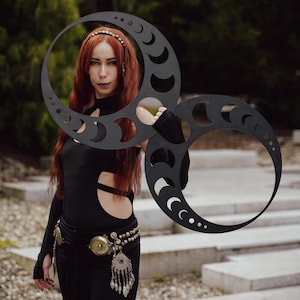 May include: A woman in a black outfit holds a large, black, crescent moon-shaped prop. The prop has cut-out crescent moon shapes and dot patterns. The woman has long red hair and wears a decorative belt.