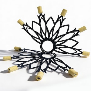 May include: A black metal fire prop featuring a symmetrical, floral-inspired cutout design with a circular centre. The structure includes eight protruding arms, each tipped with a cylindrical, light beige-coloured Kevlar wick for fire performance.