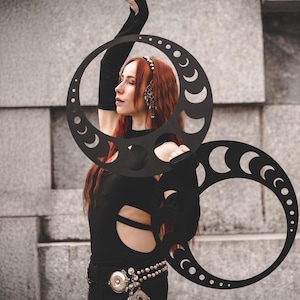May include: A woman wearing a black outfit with a cut-out design is holding two black hoops with crescent moon cut-outs. The hoops are decorated with small circles.