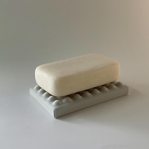 May include: A white bar of soap sits on a white soap dish with a textured surface. The soap dish has a rectangular shape with rounded edges.