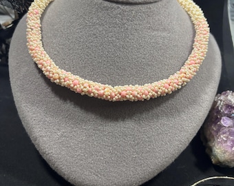 May include: A beaded necklace displayed on a gray mannequin. The necklace is a thick, circular design, featuring alternating rows of cream and light pink beads. The beads create a textured, rope-like appearance. The background is black.