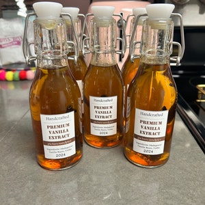 May include: Four small glass bottles filled with a brown liquid. Each bottle has a white lid and a silver clasp. The bottles are labeled "Handcrafted Premium Vanilla Extract" with the ingredients and date "2024".