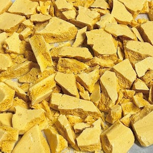 May include: A close-up of a baking sheet filled with broken pieces of honeycomb candy. The candy is a light golden yellow color and has a slightly irregular shape.