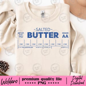 May include: A cream-coloured sweatshirt with the words "SALTED BUTTER" in blue, and a butter measurement guide. The sweatshirt also features the text "NET WT. 4 OZ. (113g)" and "GRADE AA". The bottom of the shirt has the text "Woldero | premium quality file | PNG | Digital Download".