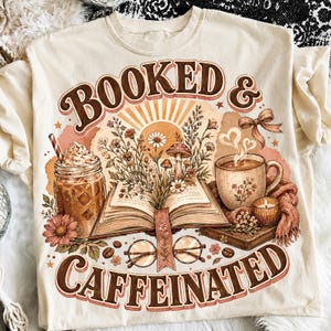 Puede incluir: Una camiseta color crema con un estampado gráfico que incluye el texto "BOOKED & CAFFEINATED". El diseño muestra un libro abierto con flores y setas, un tarro de café helado, una taza humeante, unas gafas y una pequeña vela.