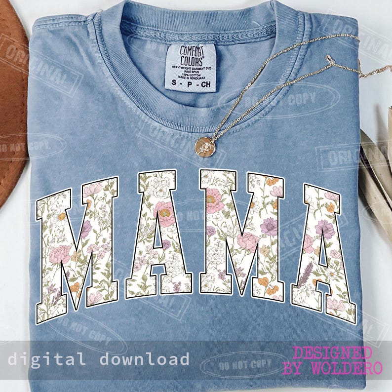 May include: A light blue t-shirt with the word "MAMA" in large, floral-patterned letters. The shirt has a crew neck and short sleeves. A gold necklace with a pendant is draped across the shirt. The shirt is labeled "COMFORT COLORS".