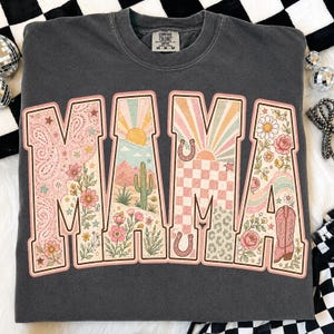 Puede incluir: Una sudadera de cuello redondo gris carb&oacute;n con la palabra MAMA en letras grandes. Cada letra contiene un dise&ntilde;o gr&aacute;fico &uacute;nico que incluye paisajes des&eacute;rticos, cactus, soles, herraduras, patrones de cuadros, flores y una bota vaquera.