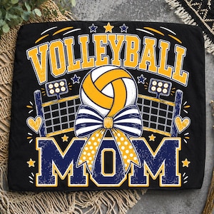 May include: A black t-shirt featuring a graphic design with the text VOLLEYBALL MOM in yellow and navy blue. The design includes a volleyball with a navy and white striped bow, a volleyball net, stadium lights, and yellow heart and star accents.
