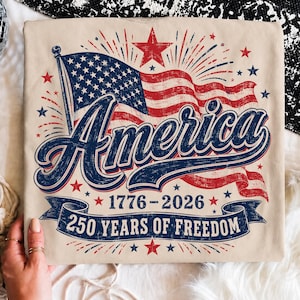 May include: A beige t-shirt featuring a graphic of a waving American flag with a large red star above it. The text reads America, 1776-2026, and 250 Years of Freedom. The design includes blue and red fireworks, stars, and a blue banner.