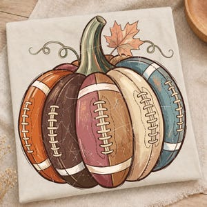 May include: A light beige t-shirt featuring a graphic of a pumpkin constructed from segments that resemble American footballs in shades of orange, brown, maroon, cream, and teal, topped with a green stem and a single orange maple leaf.