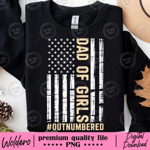 May include: Black sweatshirt featuring a distressed American flag design with the text "DAD OF GIRLS" in gold and the hashtag "#OUTNUMBERED". The design includes white stars and stripes.