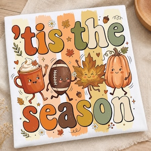 Puede incluir: Una camiseta blanca con el texto 'tis the season' en letras redondeadas y coloridas. El diseño incluye personajes de dibujos animados de una taza con nata, un balón de fútbol americano, una hoja de arce y una calabaza con iconos otoñales.