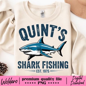 Puede incluir: Sudadera de color crema con un gráfico de tiburón azul y el texto "QUINT'S SHARK FISHING EST. 1975". El tiburón tiene detalles en blanco y negro. La sudadera es de estilo casual.