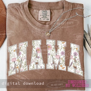 May include: A brown t-shirt with the word "MAMA" in large, white, floral-patterned letters. A silver necklace with a pendant rests on the shirt. The shirt has a vintage look and feel.