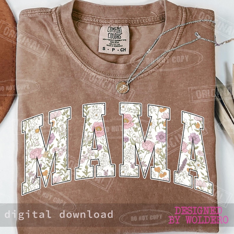 May include: A brown t-shirt with the word "MAMA" in large, white, floral-patterned letters. A silver necklace with a pendant rests on the shirt. The shirt has a vintage look and feel.