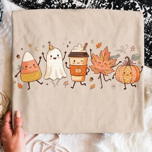 Op de afbeelding: Een beige t-shirt met een kleurrijke grafische print van vijf stripfiguren: een candy corn, een geest met een feesthoedje, een koffiebeker met slagroom, een esdoornblad en een pompoen, allemaal met lachende gezichten en dunne ledematen.