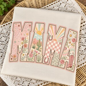 Puede incluir: Una camiseta de color crema con la palabra MAMA en letras grandes de bloque. Cada letra contiene un dise&ntilde;o gr&aacute;fico diferente, incluyendo patrones de cachemira, un paisaje des&eacute;rtico con un cactus, un sol, un tablero de ajedrez y motivos florales.