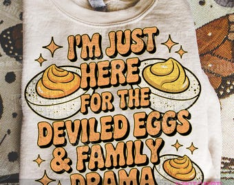 May include: A beige sweatshirt with the text "I'M JUST HERE FOR THE DEVILED EGGS & FAMILY DRAMA" in orange lettering. The design includes illustrations of deviled eggs and star shapes.