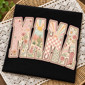 Puede incluir: Una camiseta negra con la palabra MAMA en letras grandes de bloque. Cada letra contiene un dise&ntilde;o gr&aacute;fico diferente que incluye patrones de cachemira, paisajes des&eacute;rticos con cactus, rayos de sol, cuadros, herraduras y motivos florales.