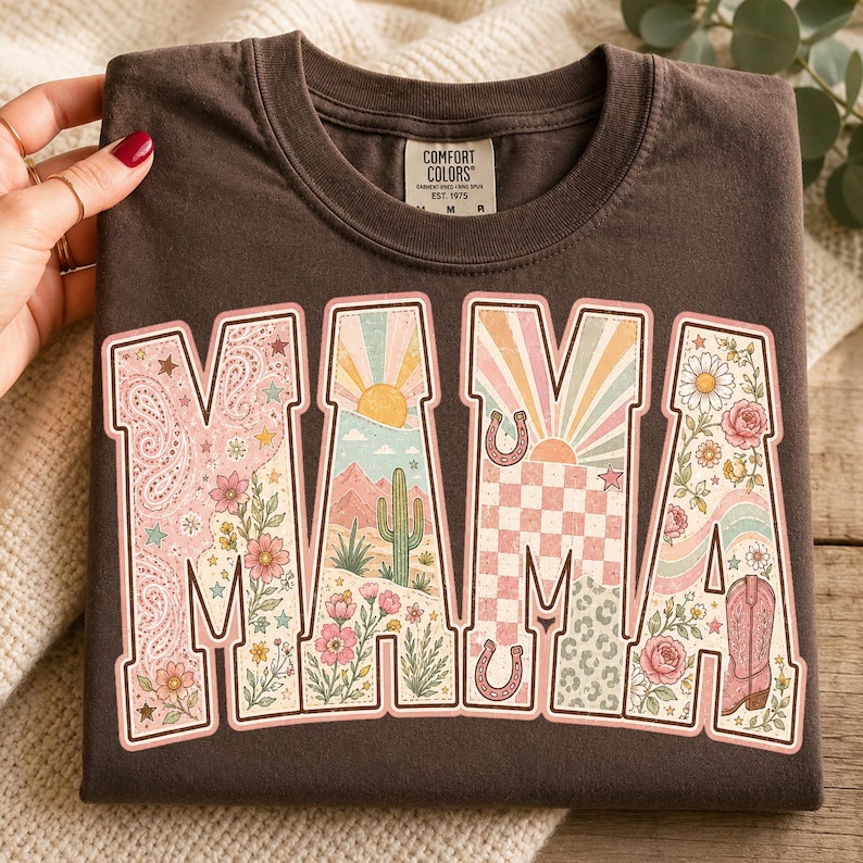 Puede incluir: Una camiseta de manga corta de color marr&oacute;n oscuro con cuello redondo y etiqueta de la marca Comfort Colors. El frente presenta la palabra MAMA en letras grandes, cada una rellena con ilustraciones coloridas que incluyen cachemira, un paisaje des&eacute;rtico, un sol, un patr&oacute;n de cuadros, flores, herraduras y una bota vaquera.
