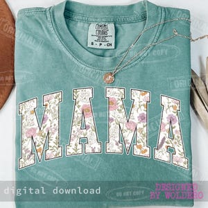May include: A teal Comfort Colors t-shirt with the word "MAMA" in large, floral-patterned letters across the front. A delicate necklace with a pendant rests on the shirt. The words "digital download" and "designed by woldero" are also visible.