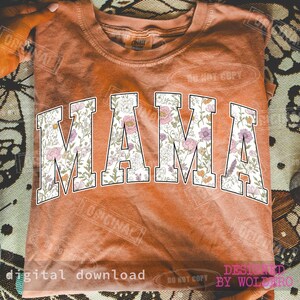 May include: A burnt orange t-shirt with the word "MAMA" in large, white, block letters. The letters are filled with a floral pattern in shades of pink, purple, and green. The shirt is folded.