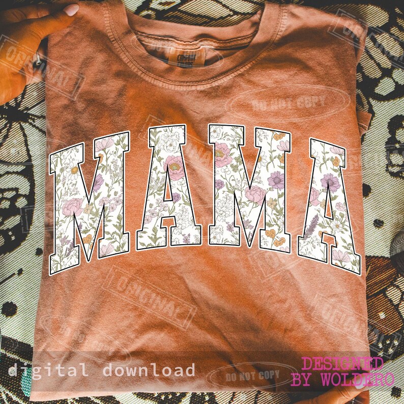 May include: A burnt orange t-shirt with the word "MAMA" in large, white, block letters. The letters are filled with a floral pattern in shades of pink, purple, and green. The shirt is folded.