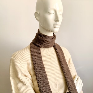 May include: A brown knitted scarf on a white mannequin. The scarf is long and thin, with a simple knit pattern.