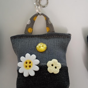 May include: A denim-style keychain in the shape of a small tote bag. The bag features a gray and black color scheme, with a white daisy and a yellow button as embellishments. The handle is gray with yellow polka dots.