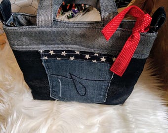 May include: A denim tote bag with a red and white polka dot ribbon. The bag has a large pocket on the front with a white star pattern and a blue denim patch. The bag is sitting on a white fluffy surface.
