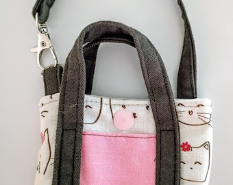 May include: Small tote bag with a cat pattern. The bag features a pink pocket, dark gray handles, and a matching strap with a metal clasp. The main fabric is white with repeating cat faces.
