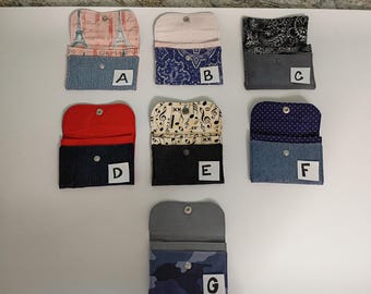 May include: A collection of seven small, open wallets with snap closures. The wallets feature various patterns and colors, including denim, paisley, musical notes, and Eiffel Tower prints. Each wallet has a white label with a letter.