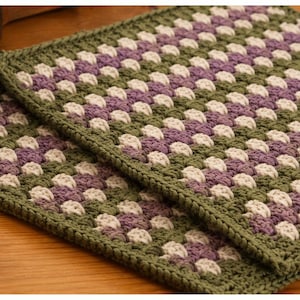 Könnte beinhalten: Zwei quadratische Häkel-Topflappen mit einem Granny-Square-Muster in den abwechselnden Farben Olivgrün, sanftes Lavendel und Creme. Jeder Topflappen ist mit einem einfarbigen olivgrünen Häkelrand an allen vier Kanten versehen.