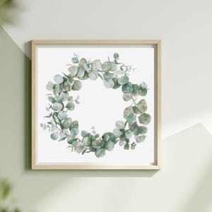 Puede incluir: Una impresión enmarcada con una corona de eucalipto en acuarela. La corona está compuesta por hojas verdes y verde grisáceo dispuestas en un patrón circular sobre un fondo blanco. El marco es de color madera clara.