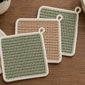 May include: Three square crochet potholders featuring a textured stitch pattern. Two potholders are sage green and one is beige, each finished with a cream-colored border and a small loop at the corner for hanging. The items are arranged in a stack.