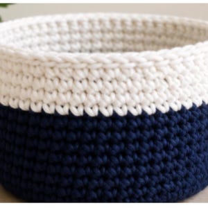 May include: A round, hand-crocheted storage basket featuring a two-tone design. The upper portion is crafted from off-white yarn, while the lower section is made from deep navy blue yarn. The basket has a thick, textured stitch pattern throughout.